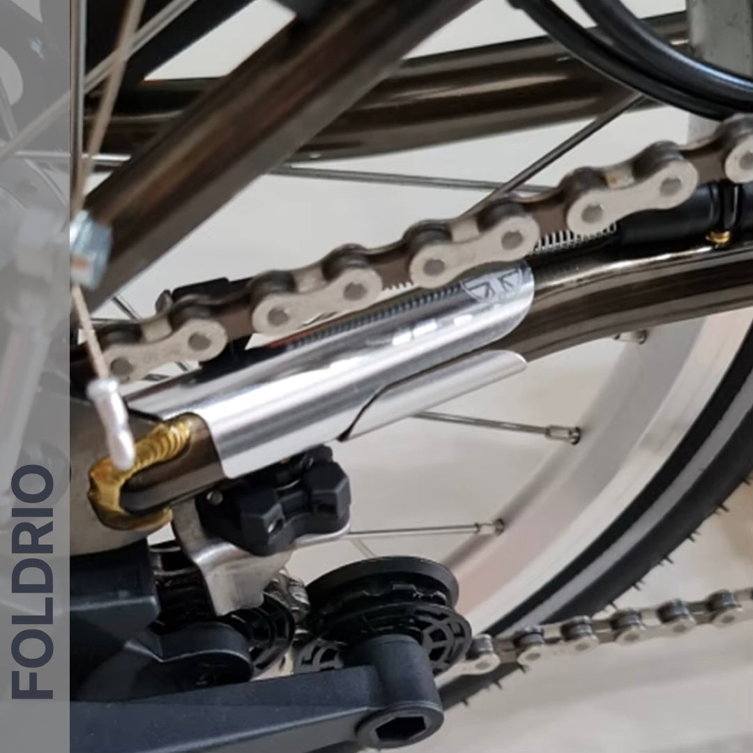 Close-up of a bicycle's rear wheel showing the silver chain tensioner and FOLDRIO Brompton Frame Protector Set with British Flag. The bike frame, spokes, and vertical "FOLDRIO" branding are visible.
