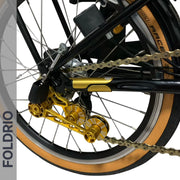 Close-up of a bicycle’s rear wheel with gold derailleur, tan sidewall tire, gold bike chain, and black frame featuring the FOLDRIO Brompton Frame Protector Set with British Flag. The FOLDRIO logo appears vertically on the left.