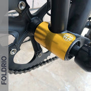 Close-up of a bicycle crankset with a gold bottom bracket and silver emblem, plus part of the chainring and pedal. A FOLDRIO Brompton Frame Protector Set with British Flag adds durability. The word FOLDRIO appears along the left edge.