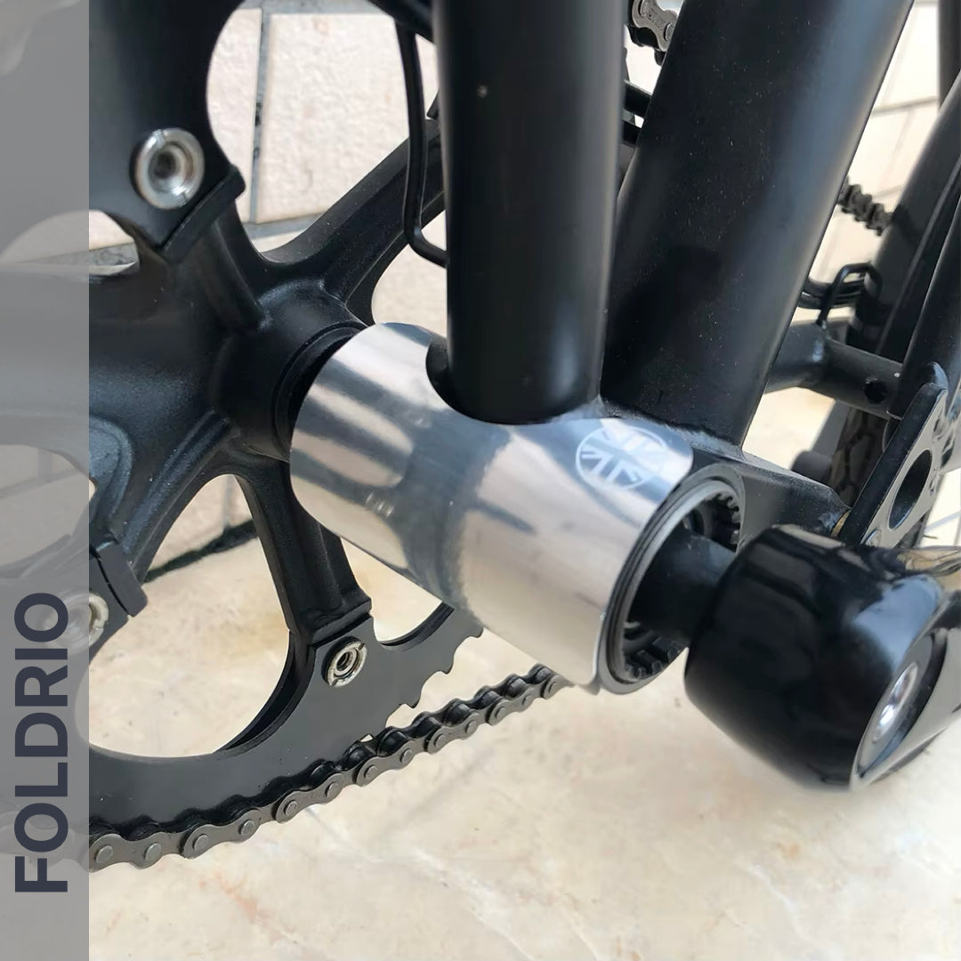 Close-up of a bicycle’s bottom bracket area featuring the crankset, chainring, metallic folding hinge with black knob, and a FOLDRIO Brompton Frame Protector Set with British Flag displayed vertically on the left side.