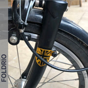 Close-up of a black bicycle fork with FOLDRIO's Brompton Frame Protector Set with British Flag emblem. Part of the front wheel, brake, and cable is visible. “FOLDRIO” appears vertically on a grey bar to the left.