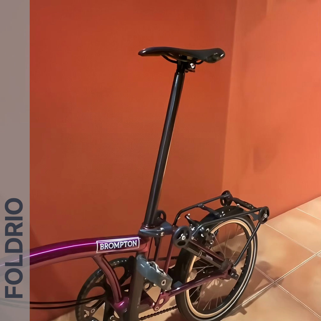 A partially folded Brompton with a purple frame and a Brompton Titanium Seatpost – Lightweight Upgrade (31.8mm, 560mm or 580mm) stands indoors by an orange wall. "FOLDRIO" appears vertically on the left edge of the image.