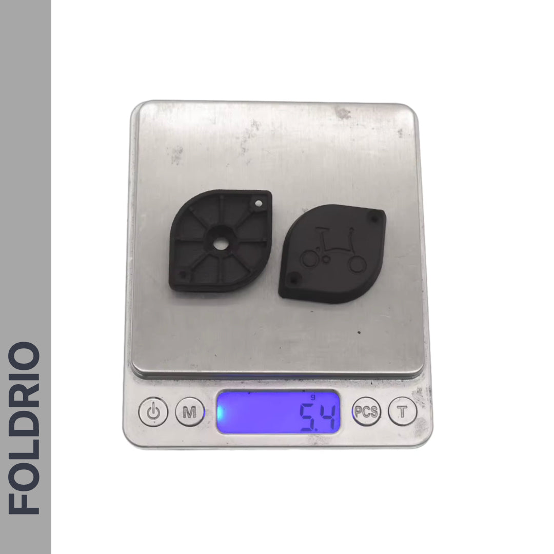 Two small black plastic parts are shown on a scale reading 5.4g. The left reveals its internal design; the right features a bicycle engraving and secretly holds an Apple AirTag. "FOLDRIO" appears vertically. Product: Brompton AirTag Saddle Mount – Hidden Tracker Holder.