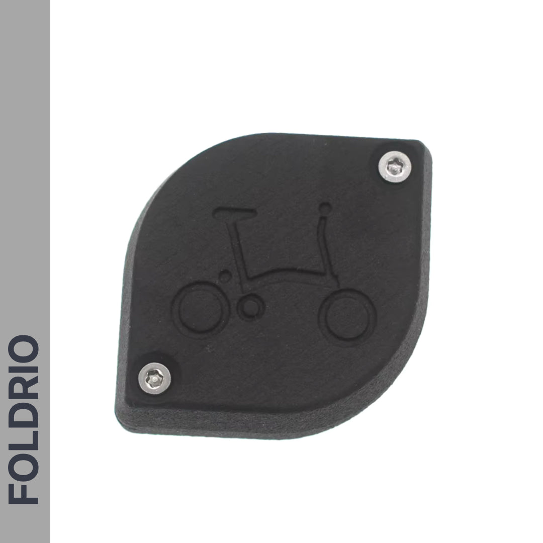 The Brompton AirTag Saddle Mount – Hidden Tracker Holder is a discreet black plastic cover with two screws and a folding bicycle outline, designed to hide your Apple AirTag for anti-theft bike tracking. FOLDRIO is displayed vertically on a gray strip.