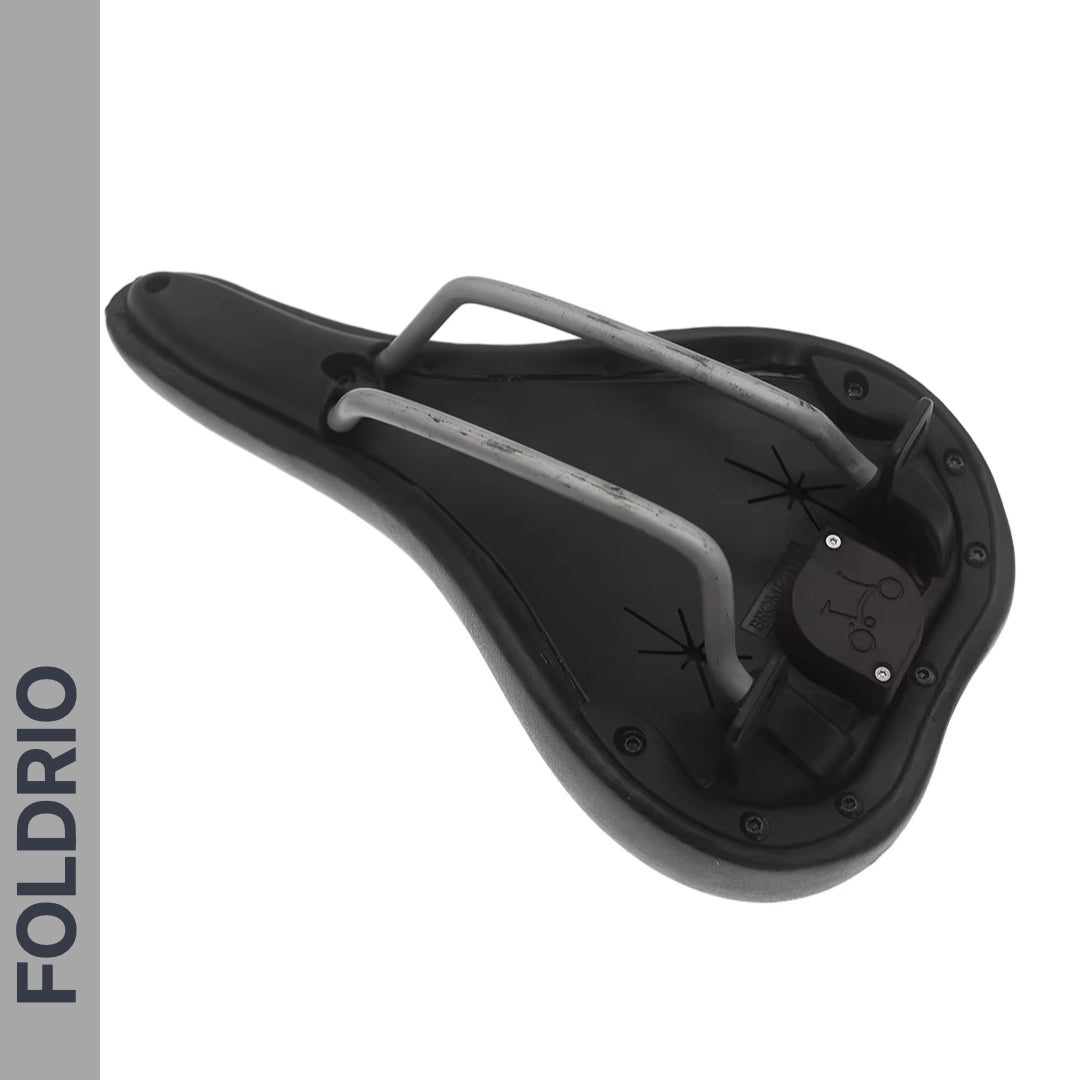 A black Brompton AirTag Saddle Mount – Hidden Tracker Holder is shown from the underside, emphasizing metal rails, mounting points, and a concealed AirTag slot. At left, a vertical gray bar displays "FOLDRIO" in bold dark letters.