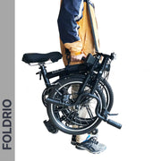 A person uses the Brompton Carry Handle & Shoulder Strap to easily hold a folded black bike by its frame. The handle and strap make carrying your compact bicycle simple and convenient.