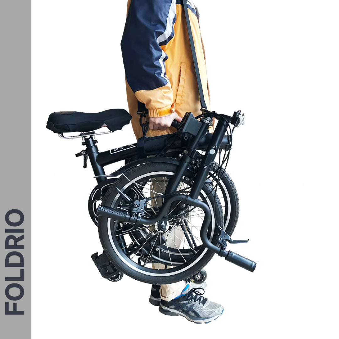 A person uses the Brompton Carry Handle & Shoulder Strap to easily hold a folded black bike by its frame. The handle and strap make carrying your compact bicycle simple and convenient.