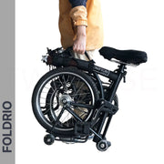 A person uses the Brompton Carry Handle & Shoulder Strap – Folded Bike Carrier to hold a folded black bicycle upright. The jacket is tan and navy, and "FOLDRIO" appears vertically on the left side.