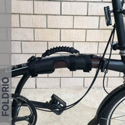 Close-up of a black folding bicycle with a Brompton Carry Handle & Shoulder Strap – Folded Bike Carrier attached to the frame, in front of a beige brick wall. "FOLDRIO" appears vertically on the left, emphasizing this innovative accessory.