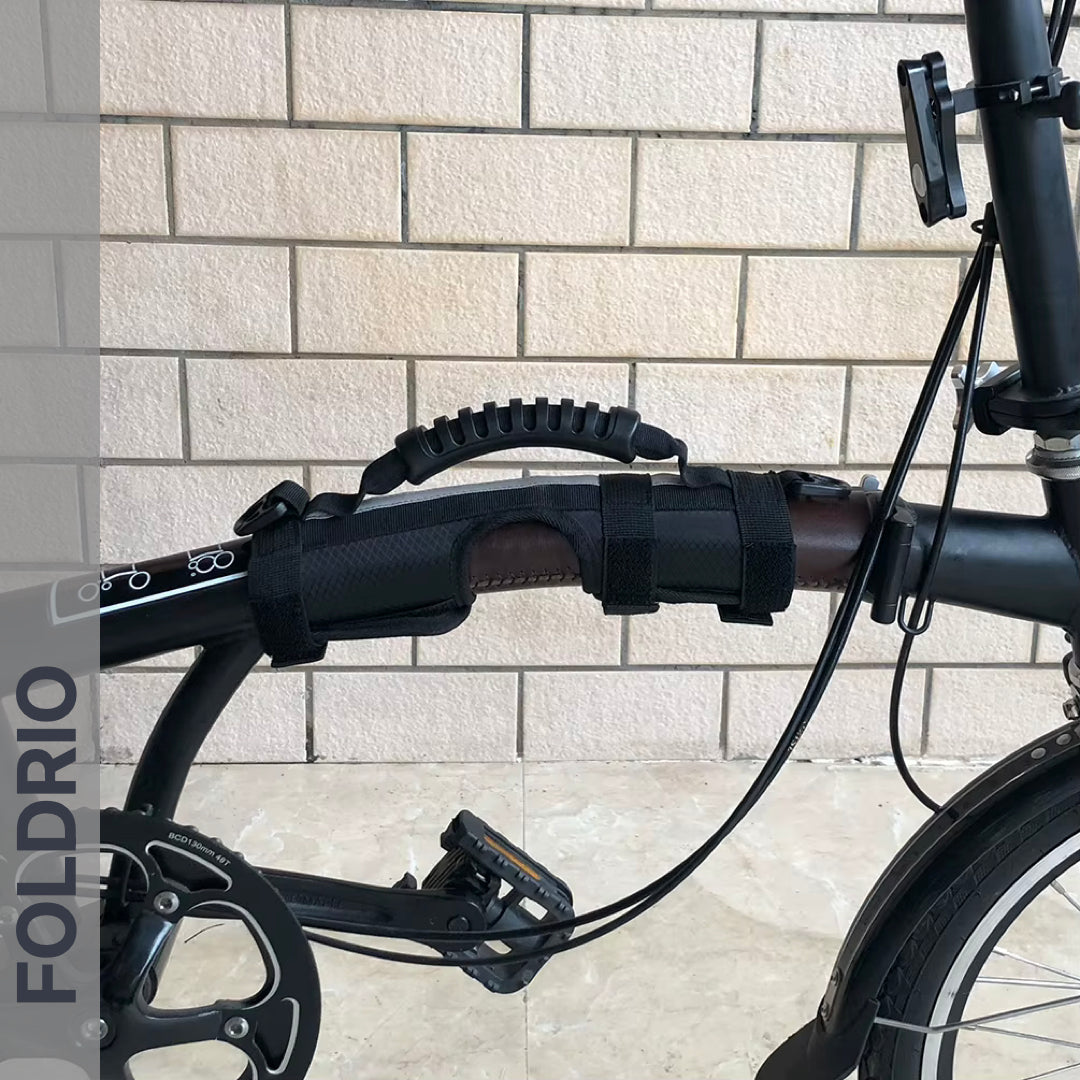 Close-up of a black folding bicycle with a Brompton Carry Handle & Shoulder Strap – Folded Bike Carrier attached to the frame, in front of a beige brick wall. "FOLDRIO" appears vertically on the left, emphasizing this innovative accessory.