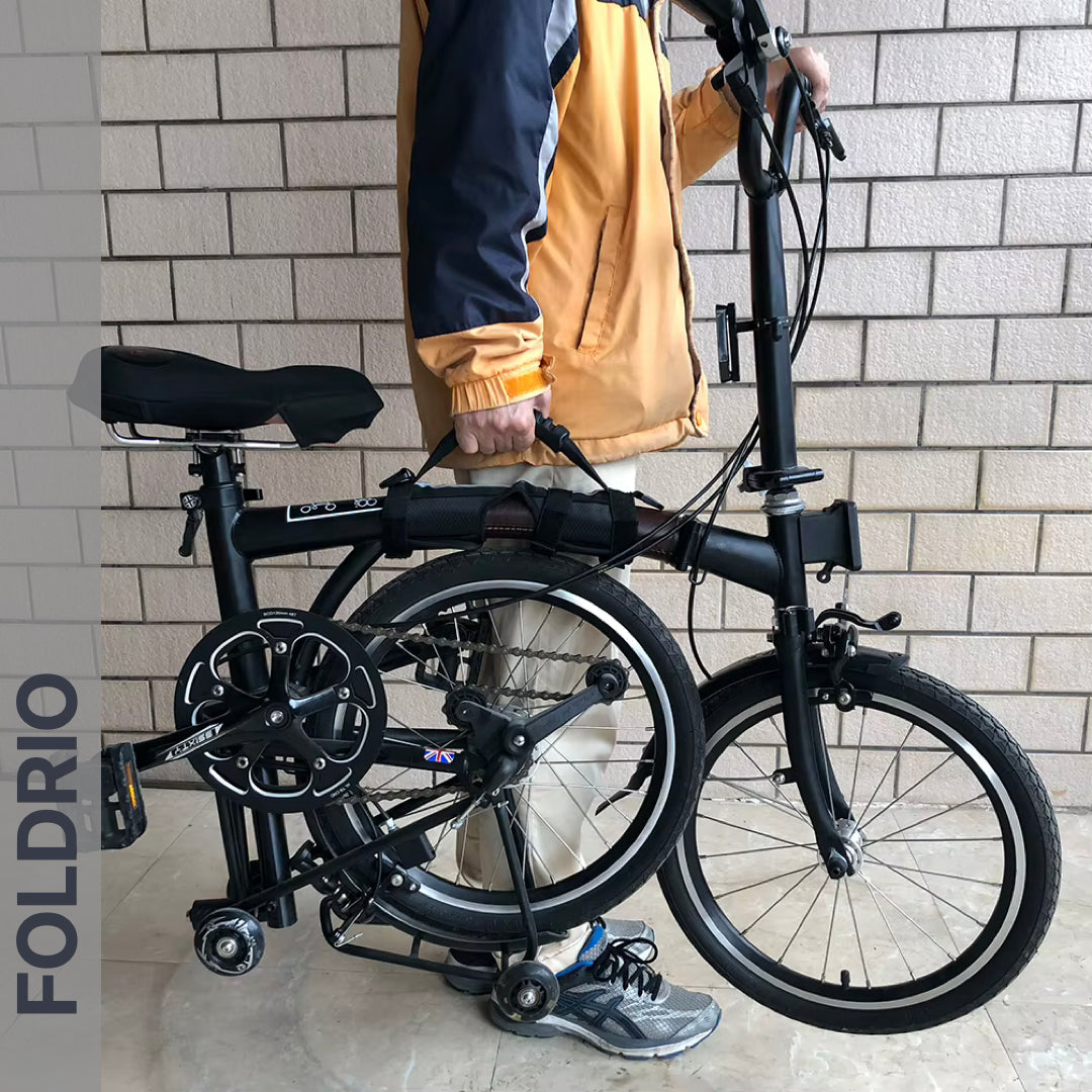 A person in a yellow and blue jacket stands holding a black folding bike fitted with the Brompton Carry Handle & Shoulder Strap – Folded Bike Carrier. The scene is set against a wall of light-colored bricks.
