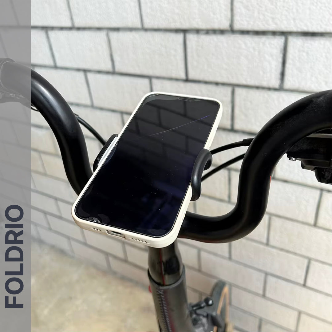 A Brompton Phone Mount – Lightweight, No Case Needed by FOLDRIO securely holds a smartphone on bicycle handlebars in front of a light-colored brick wall. The word FOLDRIO appears vertically on the left side of the image.