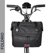 A black FOLDRIO Brompton Roll-Top Front Bag (27.5L) with buckle closures and a Union Jack patch is mounted on the handlebars via a front carrier block. The image features a gray banner with "FOLDRIO" text.