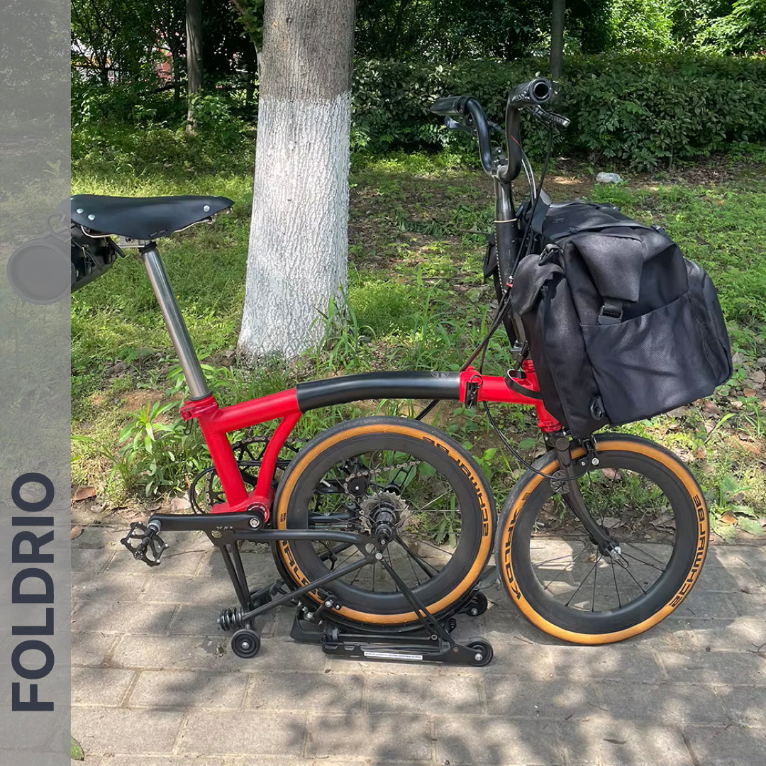 A red folding bicycle with tan tires is parked on a sidewalk near greenery, folded with a Brompton Roll-Top Front Bag – 27.5L by FOLDRIO attached to the front carrier block. A tree trunk and bushes are in the background.