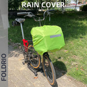 A Brompton Roll-Top Front Bag – 27.5L by FOLDRIO with a bright green rain cover is attached to a folding bike's front carrier block, parked on a sunny park path. The image displays the words RAIN COVER and FOLDRIO.