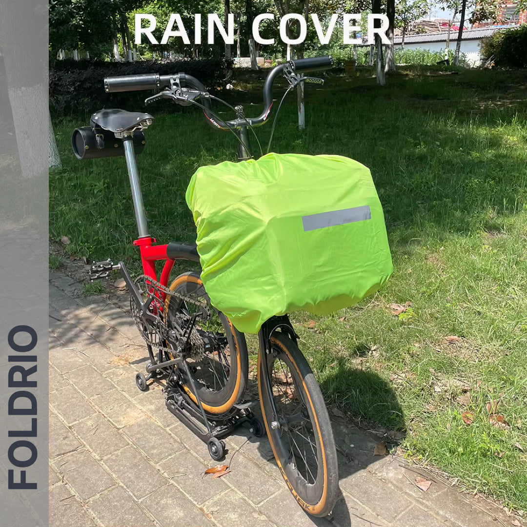 A Brompton Roll-Top Front Bag – 27.5L by FOLDRIO with a bright green rain cover is attached to a folding bike's front carrier block, parked on a sunny park path. The image displays the words RAIN COVER and FOLDRIO.
