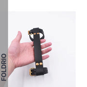 A hand holds the FOLDRIO Brompton Leather Carry Handle – Full-Grain with brass hardware, featuring adjustable buckle straps. The product is shown against a white background, with "FOLDRIO" displayed vertically on the left side of the image.