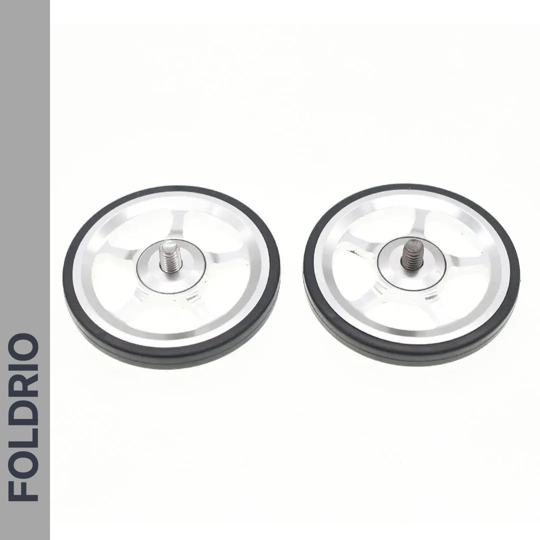 Two small silver Brompton Easy Wheels with titanium bolts, black rubber edges, and FOLDRIO branding are shown side by side on a white background, with the word FOLDRIO vertically on a gray strip to the left.