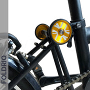 Close-up of a black bicycle frame with FOLDRIO branding, showcasing Brompton Easy Wheels Union Jack Edition with titanium bolts in gold, featuring white geometric patterns, alongside the rear wheel and chain.