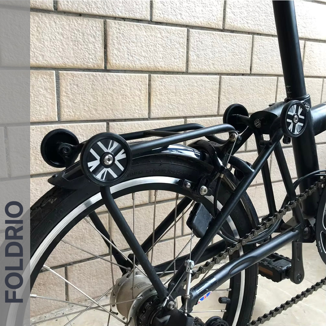 Close-up of a FOLDRIO folding bike’s rear section with Brompton Easy Wheels Union Jack Edition (titanium bolts), rear rack, chain, and small tire—all set against a brick wall backdrop.