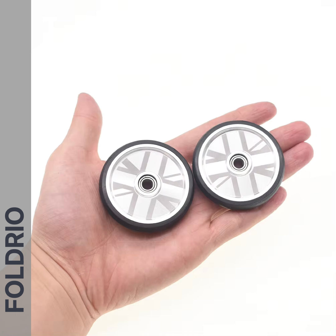 A hand holds two compact, silver and black Brompton Easy Wheels Union Jack Edition with titanium bolts by FOLDRIO; the brand name appears vertically on the left, accentuating these stylish aluminium bike wheels.
