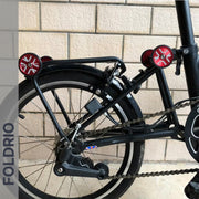 Close-up of the rear wheel and folding mechanism of a FOLDRIO Brompton Easy Wheels Union Jack Edition with titanium bolts, shown against a tiled wall. The brand name FOLDRIO appears vertically on the left.