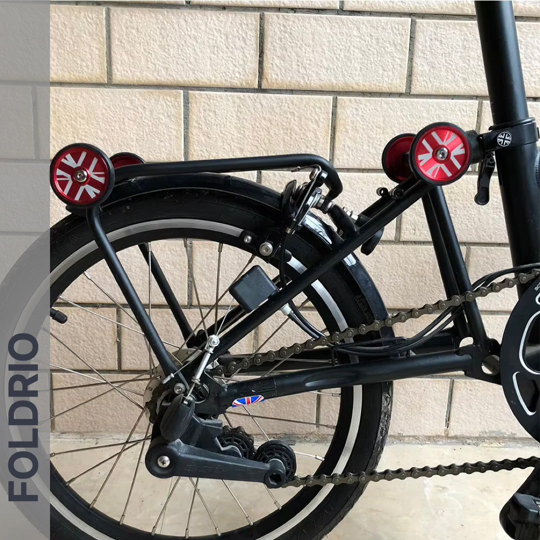 Close-up of the rear wheel and folding mechanism of a FOLDRIO Brompton Easy Wheels Union Jack Edition with titanium bolts, shown against a tiled wall. The brand name FOLDRIO appears vertically on the left.