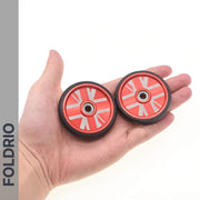 A hand holds two small red and white FOLDRIO Brompton Easy Wheels Union Jack Edition with black edges, metal bearings, and titanium bolts. The word FOLDRIO appears vertically on the left side of the image.