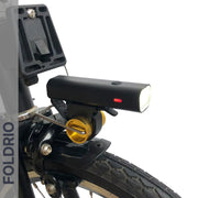 Close-up of the FOLDRIO Brompton Front Fork Light – 400 Lumen, USB-C Rechargeable, mounted above the front wheel on a black fork beside a yellow reflector and tire tread. "FOLDRIO" is displayed vertically on the left side of the image.