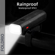 A black FOLDRIO Brompton Front Fork Light, mounted on a handlebar and covered with water droplets, shines a bright 400-lumen adjustable beam. Text reads "Rainproof Waterproof IPX3." The light is USB-C rechargeable.
