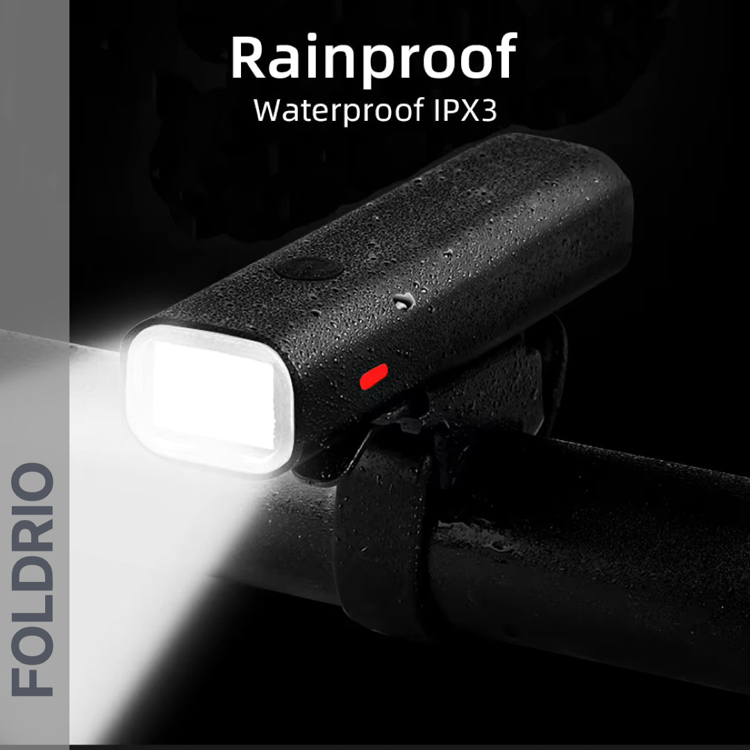 A black FOLDRIO Brompton Front Fork Light, mounted on a handlebar and covered with water droplets, shines a bright 400-lumen adjustable beam. Text reads "Rainproof Waterproof IPX3." The light is USB-C rechargeable.