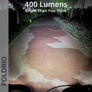 A bike path glows under the Brompton Front Fork Light – 400 Lumen by FOLDRIO, revealing the way and nearby bushes. Text: "400 Lumens, Brighter Than You Think." Left side: FOLDRIO. Highlight: adjustable beam for versatility. USB-C rechargeable.