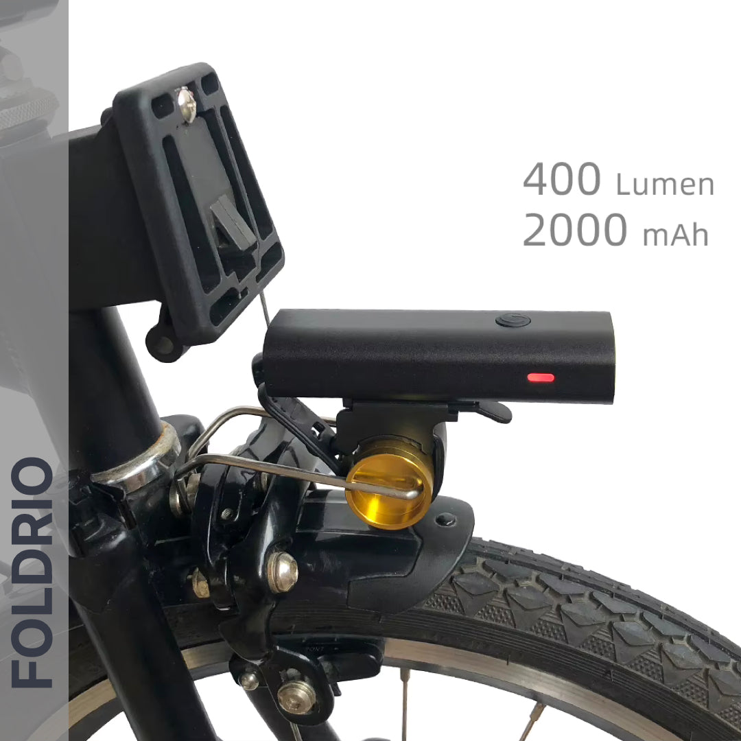 Close-up of the FOLDRIO Brompton Front Fork Light (400 lumens) in black, mounted above the front tire. Features an adjustable beam, 2000 mAh capacity, USB-C rechargeability, and FOLDRIO branding vertically on the left side.