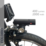 Close-up of the FOLDRIO Brompton Front Fork Light mounted above the wheel, with red indicator and adjustable 400-lumen beam. USB-C rechargeable, 2000mAh. "FOLDRIO" appears vertically on the left.
