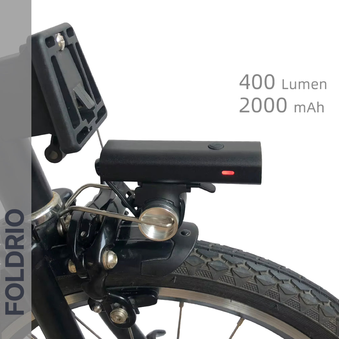 Close-up of the FOLDRIO Brompton Front Fork Light mounted above the wheel, with red indicator and adjustable 400-lumen beam. USB-C rechargeable, 2000mAh. "FOLDRIO" appears vertically on the left.