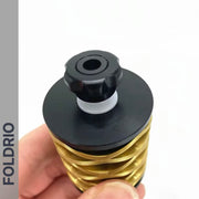 A hand holds the FOLDRIO Brompton Rear Shock Absorber, a gold and black cylindrical object with a spiral design and black knob. The word FOLDRIO appears vertically on the left side of the image.