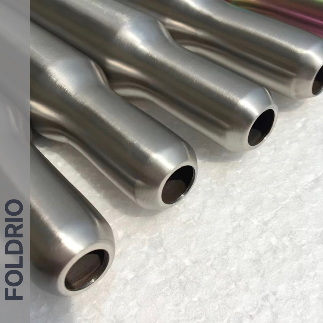 Four metallic, cylindrical Brompton Titanium Seatpost – Multicolour Edition tubes by FOLDRIO are arranged parallel on a textured white background. The FOLDRIO brand name is vertically printed along the left side of the image.