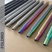 Several Brompton Titanium Seatpost – Multicolour Edition rods from FOLDRIO—finished in black, silver, and iridescent rainbow—are neatly arranged on a cardboard surface. The brand name FOLDRIO appears vertically on the left side.