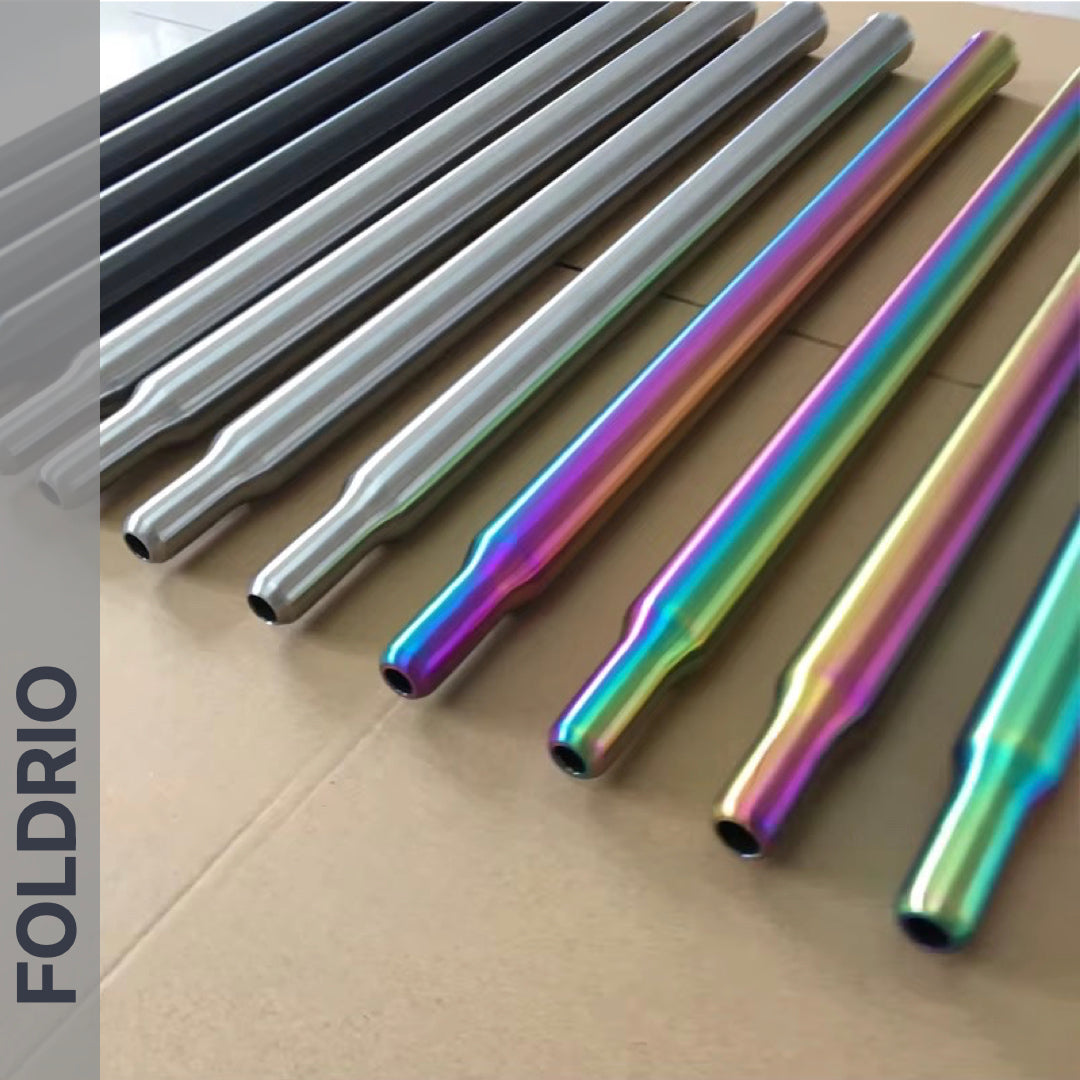 Several Brompton Titanium Seatpost – Multicolour Edition rods from FOLDRIO—finished in black, silver, and iridescent rainbow—are neatly arranged on a cardboard surface. The brand name FOLDRIO appears vertically on the left side.