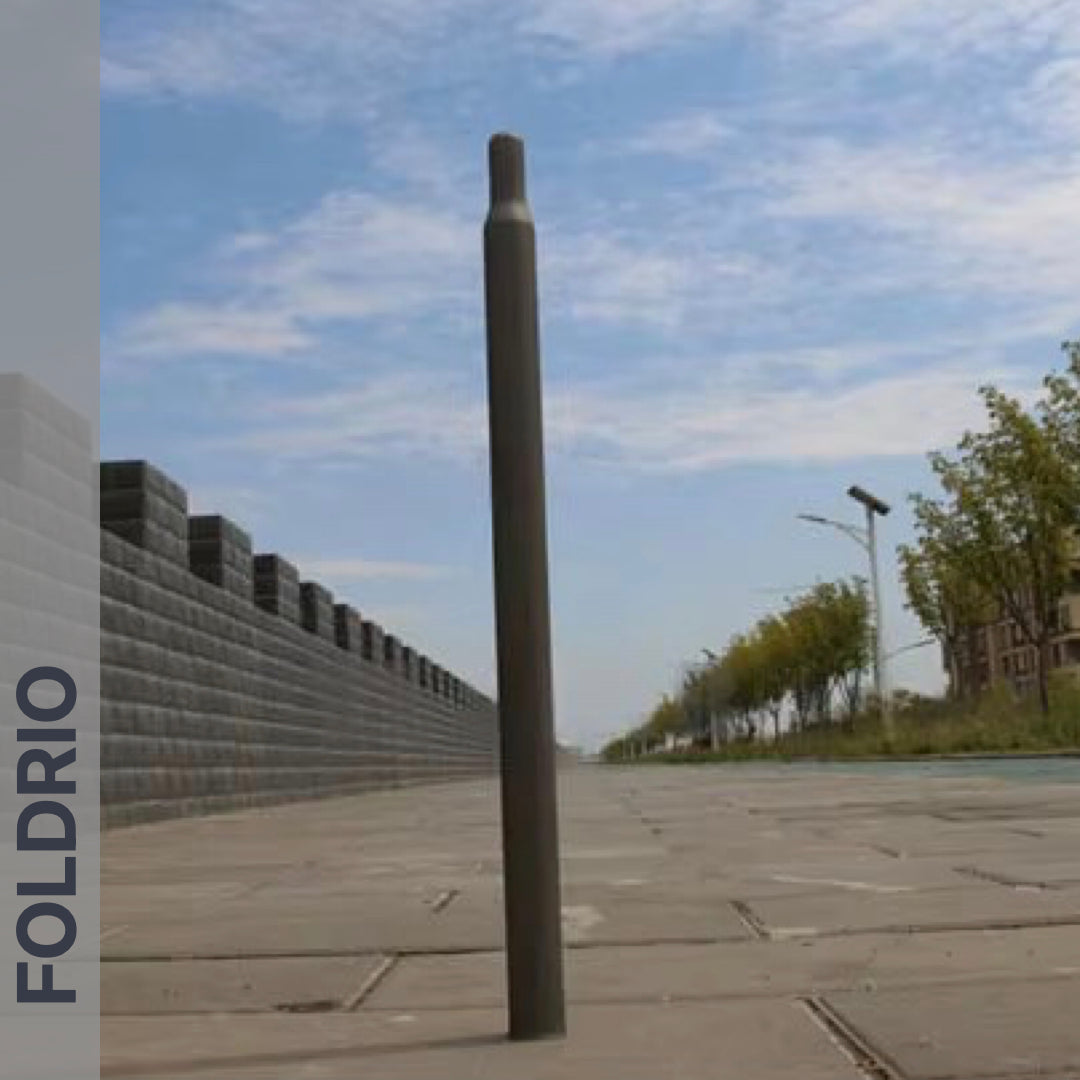 A Brompton Titanium Seatpost – Multicolour Edition by FOLDRIO stands upright on a paved sidewalk next to a stone wall and trees under a partly cloudy sky. The brand name FOLDRIO is vertically written on the left side.