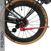 Close-up of a bicycle’s rear wheel featuring a tan Kenda tire, black Shimano derailleur, disc brake, red quick-release lever, and the FOLDRIO Brompton Kickstand – Adjustable Aluminium (A and C Line), with “FOLDRIO” vertically on the left.