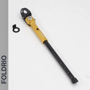 A FOLDRIO Brompton Kickstand – Carbon Fibre, P and T Line in gold and black rests on a white surface next to a small black circular part. FOLDRIO is shown vertically on a grey strip to the left.