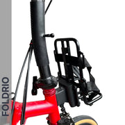 Close-up of a black FOLDRIO Brompton Front Bag Rack Holder with Elastic Straps, mounted on the front of a red Brompton folding bike. Handlebars and cables are visible against a white background.
