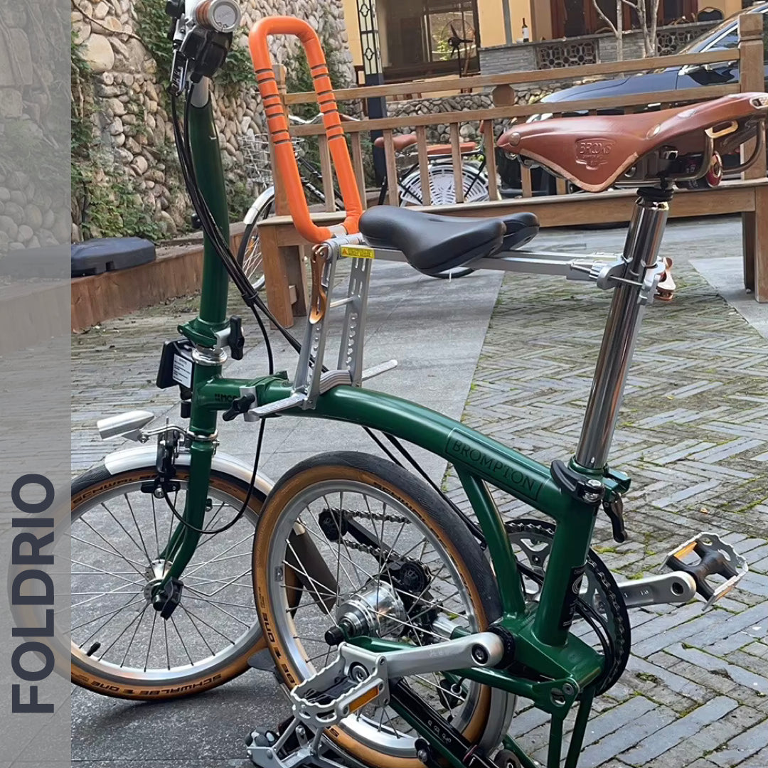 Brompton Brompton green folding bike with brown seat, orange bar, and front FOLDRIO child seat is parked by stone and wood walls | Foldrio. Foldrio