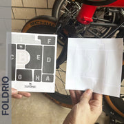 Brompton Brompton rear wheel shown as a hand holds two FOLDRIO Protective Sticker sheets, one labeled, beside the frame | Foldrio. Foldrio