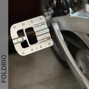 Close-up of a Brompton Foldable Flat Pedal—lightweight aluminium—mounted on a silver crank arm. A white bike frame and rear tire are in the background. The word FOLDRIO is shown vertically on the left margin.