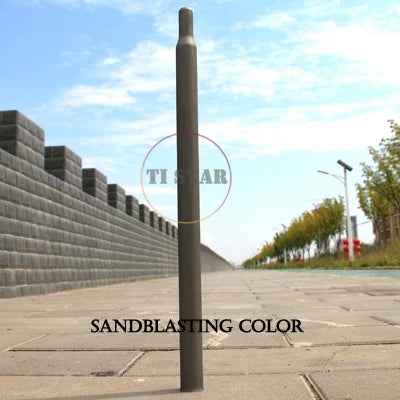 A FOLDRIO Brompton Titanium Seatpost – Multicolour Edition stands upright on a paved walkway, with a brick wall to the left and trees to the right beneath a blue sky. The words SANDBLASTING COLOR appear at the bottom.